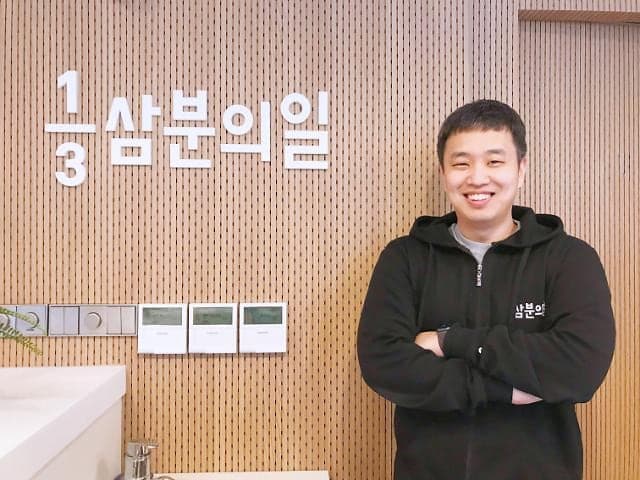 삼분의일 로고가 걸린 나무 벽 앞에서 팔짱을 끼고 웃는 전주훈 대표